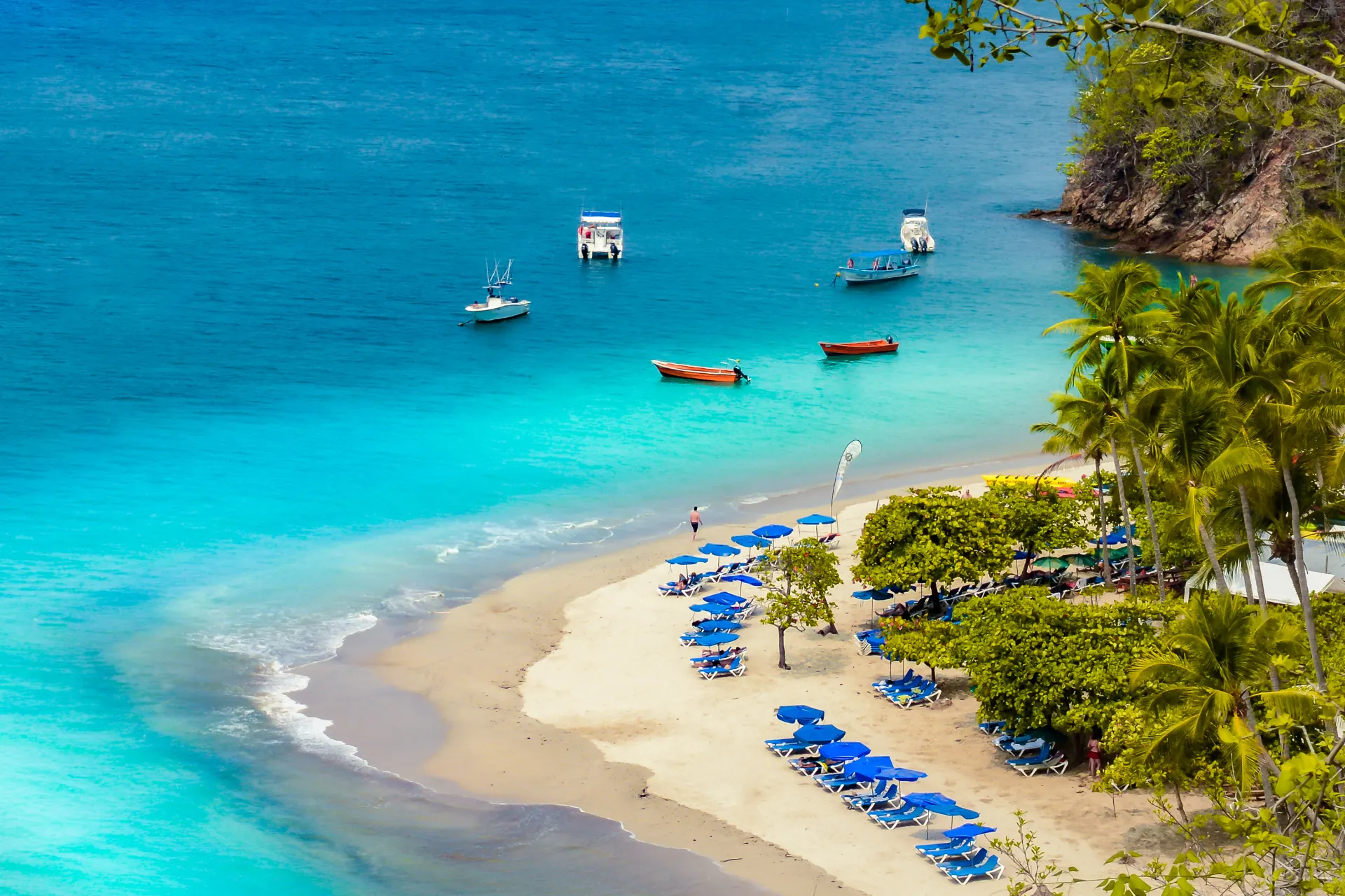 Tropisch strand met helder turquoise water en wit zand, omzoomd door palmbomen en rijen blauwe ligstoelen onder parasols. In de baai liggen kleine boten en motorbootjes op het kalme water, met een groene heuvel op de achtergrond. Ideaal voor content over exotische vakanties, strandbestemmingen, luxe resorts, zonvakanties en tropisch toerisme. Perfect voor blogs over ontspanning, duurzame reizen, idyllische kustlandschappen en tips voor strandactiviteiten.