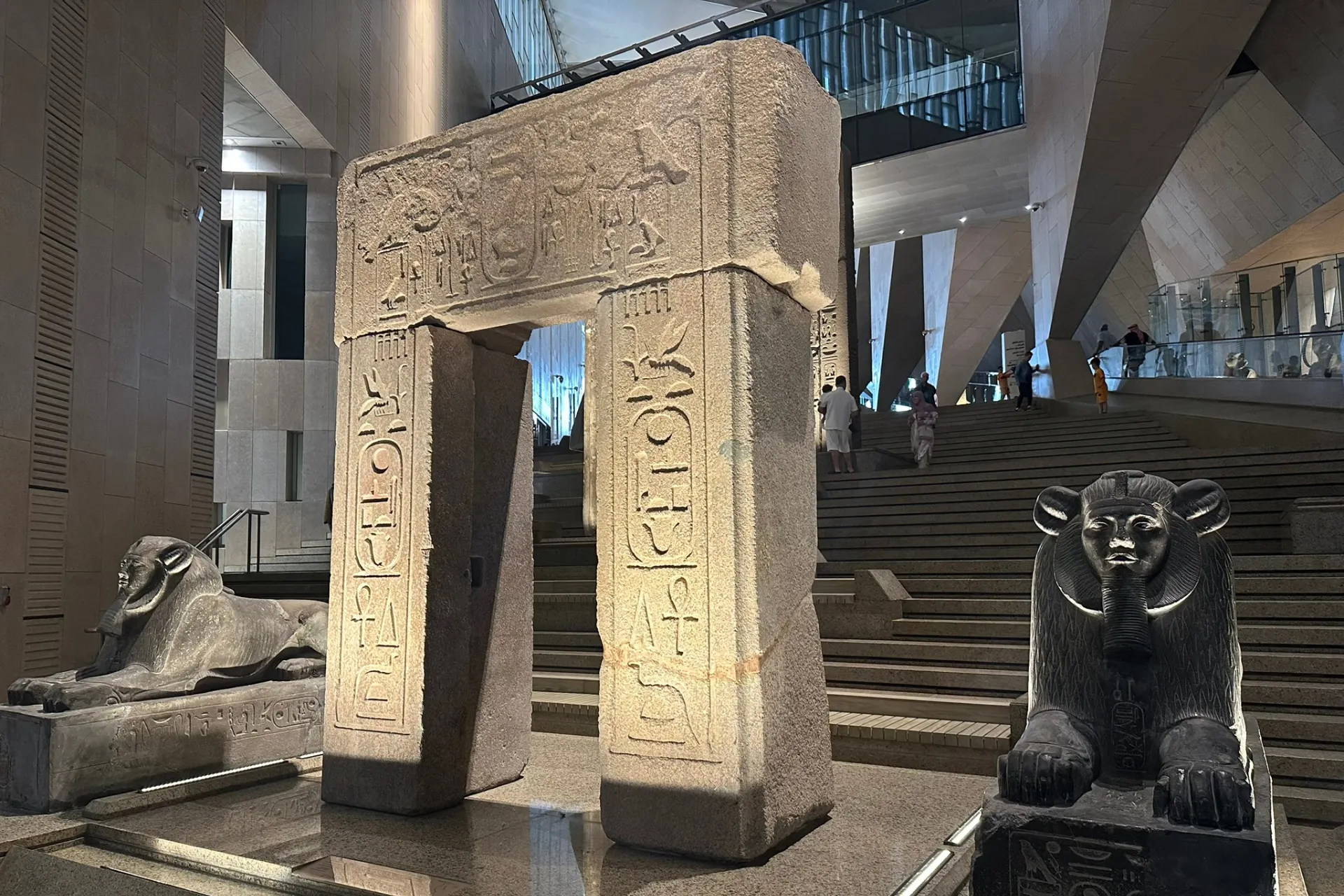 Binnen in het Egyptisch Museum met imposante stenen dierenbeelden, een sfeervol verlichte stenen poort en een trap op de achtergrond die leidt naar een hoger gelegen tentoonstellingsruimte.