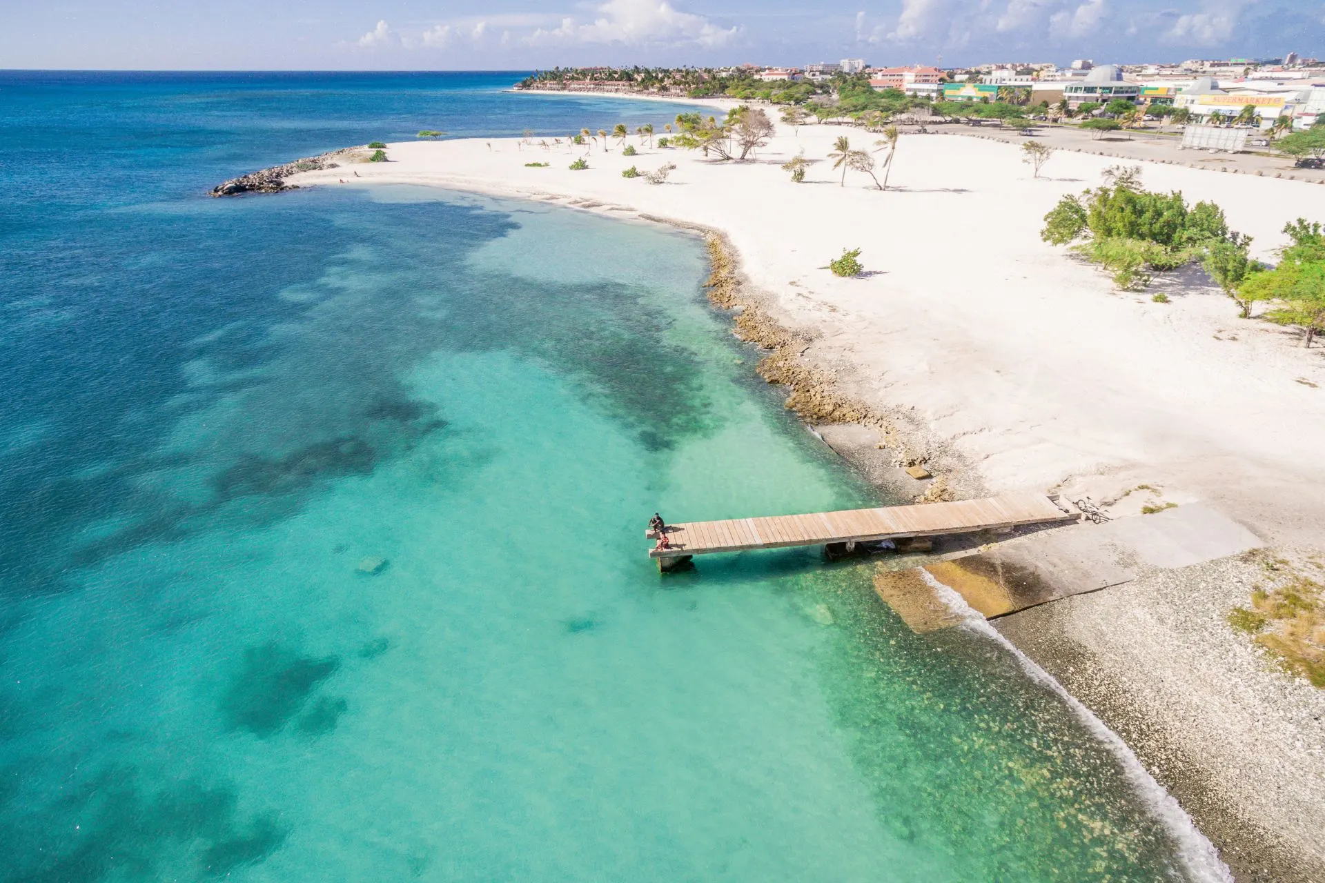 Een luchtfoto van een wit zandstrand op Aruba, omringd door helder turquoise water. Een houten pier steekt uit over het water, waarop een persoon staat te genieten van het uitzicht. In de verte zijn palmbomen, gebouwen en een rustige blauwe horizon zichtbaar, wat een serene en tropische sfeer creëert.