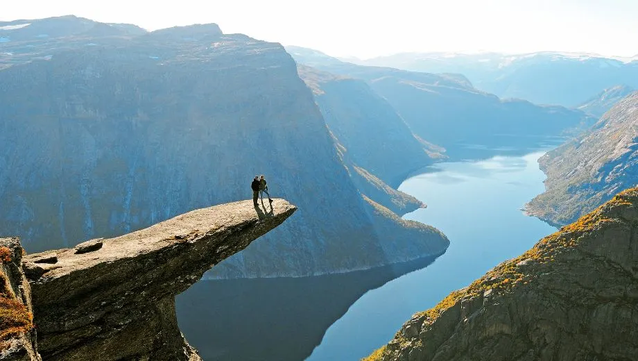 Twee wandelaars staan op de rotsformatie Trolltunga in Noorwegen, hoog boven een fjord en omringd door steile bergen.
