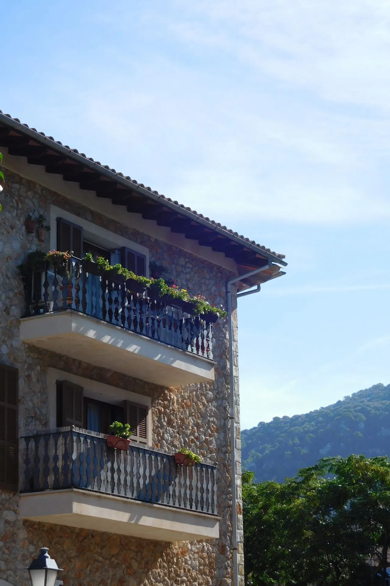 Traditioneel stenen huis met houten luiken en twee balkons vol bloemen in Valldemossa, Mallorca, met uitzicht op groene bergen onder een heldere blauwe lucht. Valldemossa is een pittoresk bergdorp in de Serra de Tramuntana, bekend om zijn charmante straatjes, historische gebouwen en mediterrane sfeer. Perfecte bestemming voor een dagtrip tijdens een vakantie op Mallorca.
