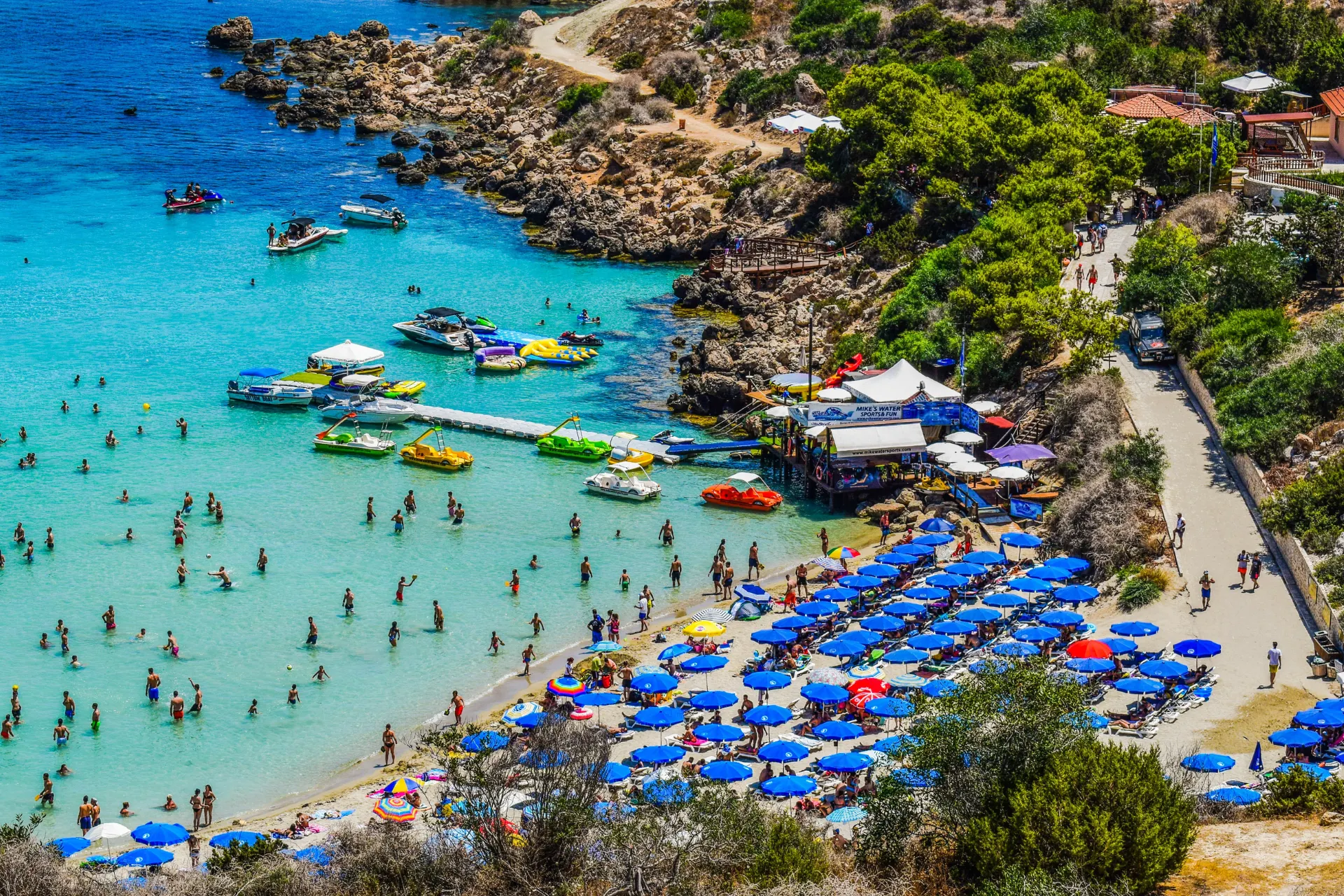 Drukke stranddag in Ayia Napa, Cyprus, gezien van bovenaf, met opvallend veel blauwe parasolletjes.