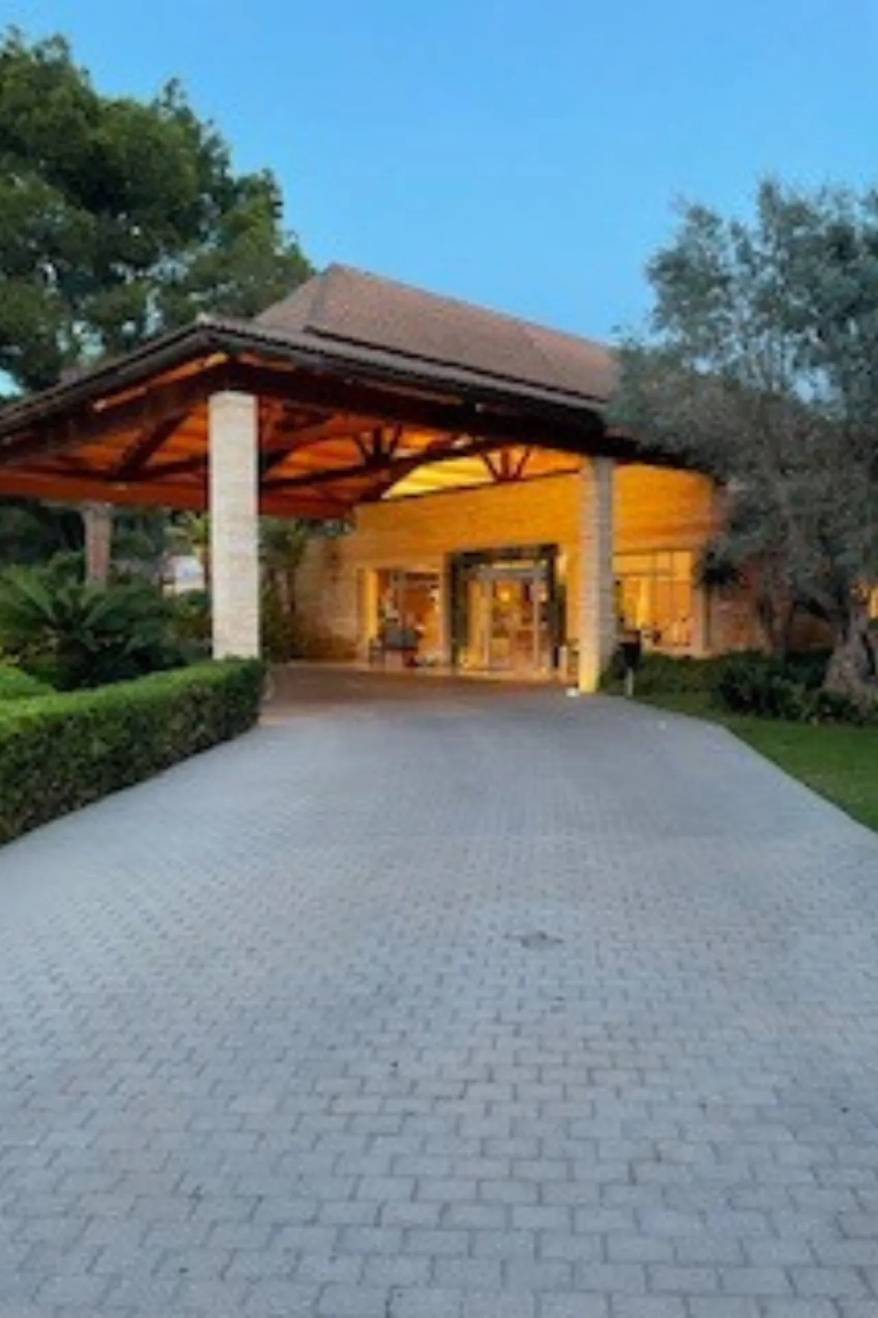 Stijlvolle hotelentree met een brede oprit van grijze klinkers, omgeven door groene struiken en bomen. Het gebouw heeft een houten dakconstructie en warme verlichting die een uitnodigende sfeer creëert bij schemering. Perfecte locatie voor een luxe verblijf, resortvakantie en ontspanning in een mediterrane omgeving met moderne faciliteiten en gastvrije ambiance.

