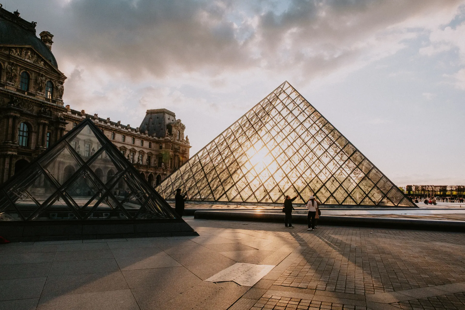 De glazen piramide van het Louvre in Parijs bij zonsondergang, met zonlicht dat door de structuur schijnt. Op de voorgrond staat een kleinere piramide, terwijl het historische museumgebouw met klassieke architectuur links zichtbaar is. Het plein is rustig met enkele bezoekers. Iconische bezienswaardigheid en cultureel symbool van Frankrijk, populair voor kunst, architectuur en fotografie.