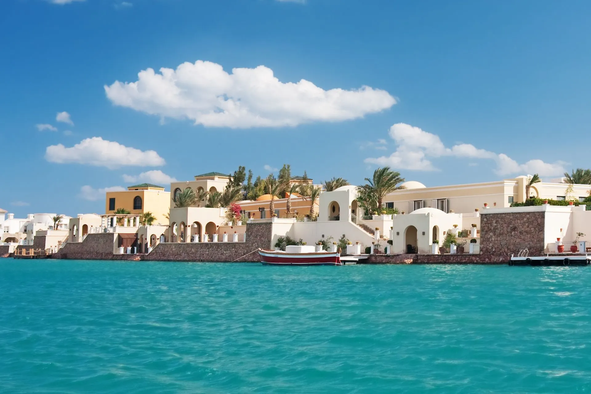 Witgekleurde villa’s en resortgebouwen langs een helderblauwe lagune in El Gouna, Egypte. De bebouwing in mediterrane en Nubische stijl ligt direct aan het water, omgeven door palmbomen en een rustige promenade. Het turquoise water en de wolkenloze lucht benadrukken de luxe en ontspannen sfeer van deze badplaats aan de Rode Zee.
