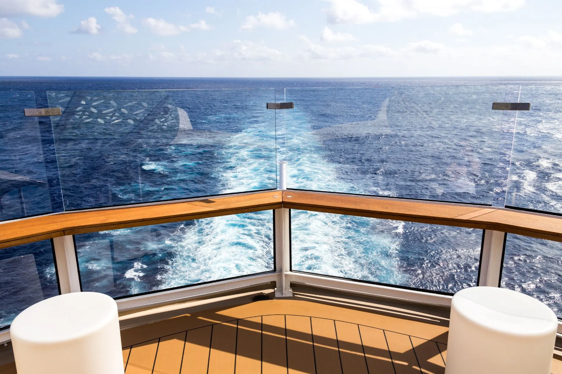 Uitzicht vanaf het dek van een cruiseschip, met een houten railing en glazen panelen. In de verte is de eindeloze blauwe oceaan te zien met witte golven die achter het schip ontstaan.