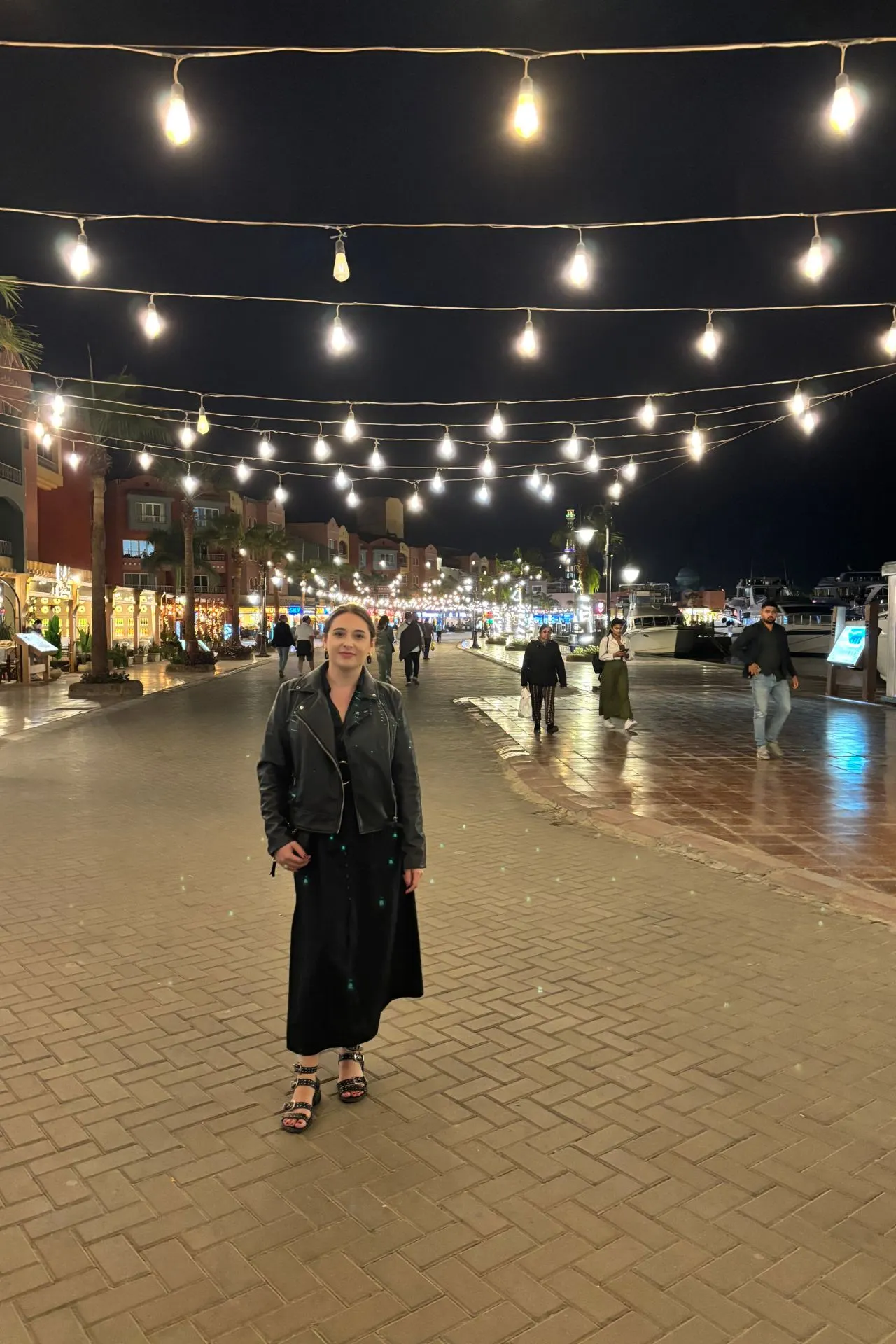 Gezellig nachtbeeld van een promenade met sfeervolle verlichting door rijen hangende lampen boven een geplaveid pad. Aan beide zijden van de straat zijn verlichte winkels, restaurants en terrassen zichtbaar, terwijl enkele mensen wandelen langs het water waar boten liggen aangemeerd. De warme lichtreflecties op de bestrating creëren een uitnodigende en levendige sfeer. Dit beeld is ideaal voor promotie van avondwandelingen, uitgaan en het bruisende nachtleven in Hurghada, Egypte.