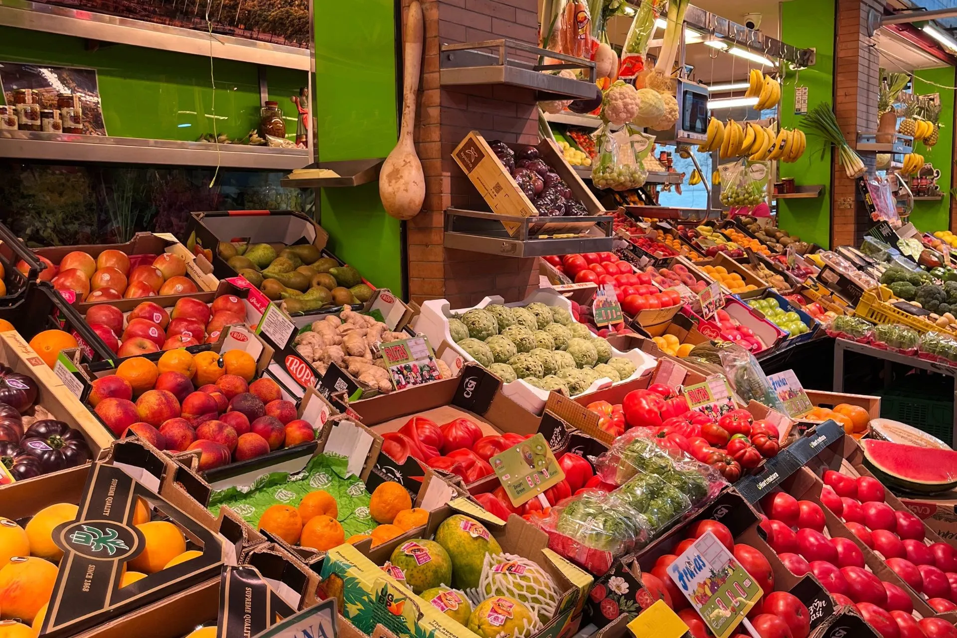 Kraam op een overdekte markt in Sevilla met kleurrijk uitgestalde groenten en fruit, waaronder tomaten, perziken, mango’s, bananen en cactusvijgen. De kraam is frisgroen aangekleed en er hangt een grote houten lepel aan de muur.

