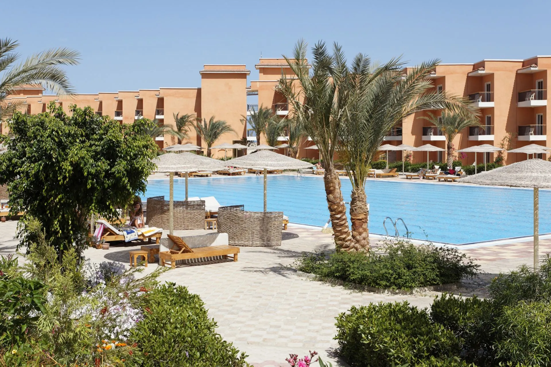 Groot buitenzwembad met ligbedden en parasols, omgeven door palmbomen en oranje resortgebouwen onder een heldere blauwe lucht.
