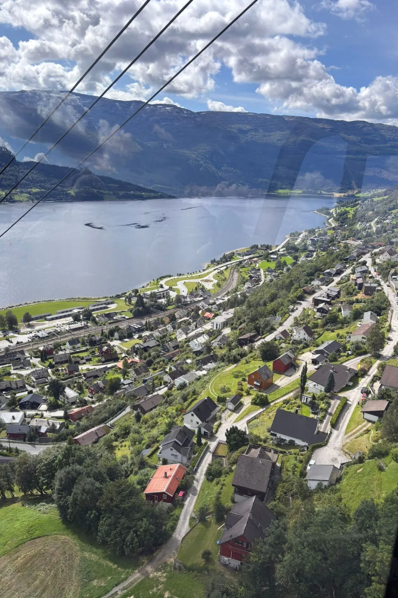 Uitzicht vanuit een hoogte over een dorp aan een groot meer, omringd door groene heuvels en bergen. Het dorp heeft verspreide huizen met rode, grijze en witte daken, kronkelende wegen en een heldere blauwe lucht met wolken erboven.