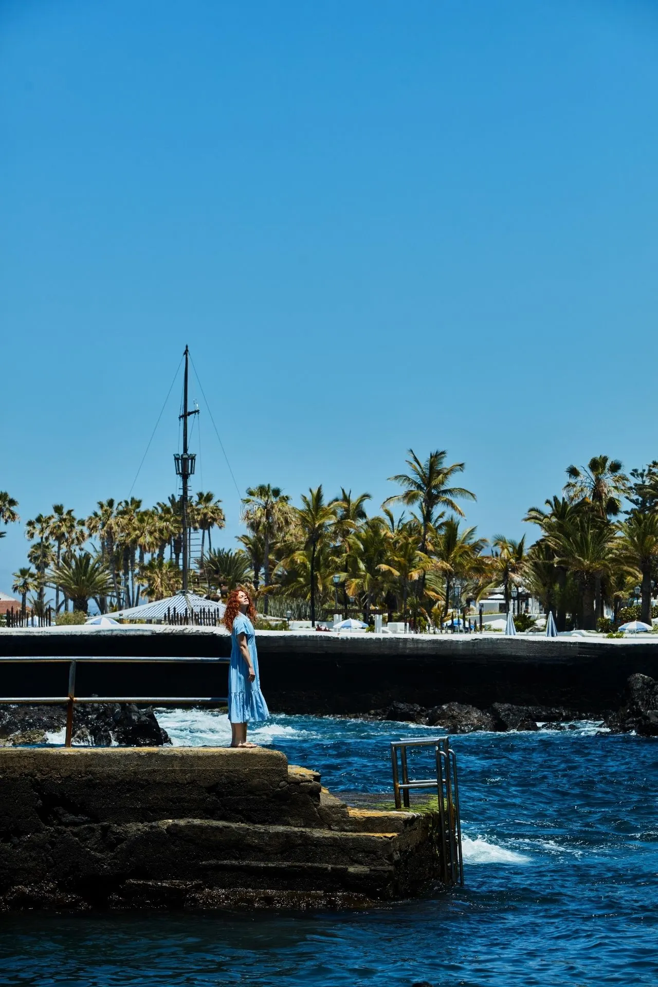 Kustlijn met blauw zeewater, palmbomen en een persoon bij de rand van een pier op Tenerife.