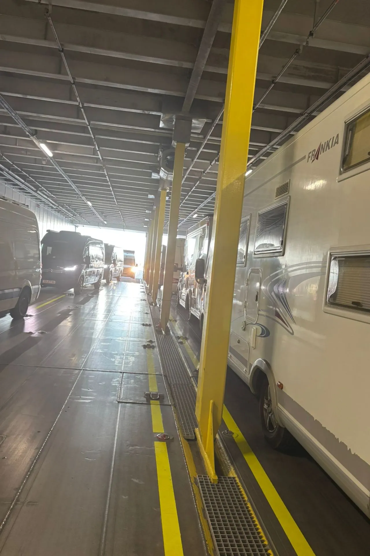 Rij campers en bestelwagens geparkeerd in een goed verlichte, overdekte ruimte met gele veiligheidsstrepen en metalen roosters op de vloer, waarschijnlijk het laaddek van een veerboot.