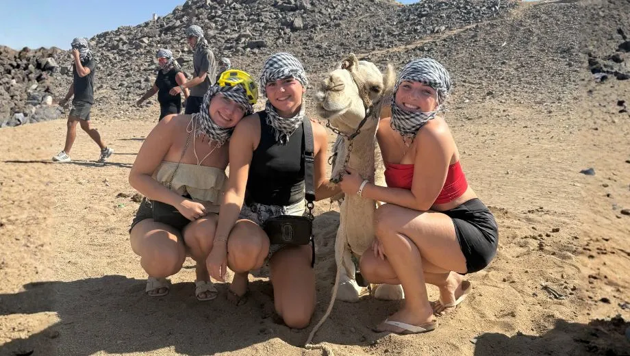 Drie personen gehurkt naast een witte kameel in een woestijnlandschap met rotsachtige heuvels op de achtergrond. Ze dragen sjaals om hun hoofd en lichte zomerkleding. Andere mensen en kamelen zijn zichtbaar in de verte.