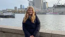 Eva Hagemeijer poseert glimlachend voor de rivier de Theems in Londen, met het iconische London Eye en moderne stadsgebouwen zichtbaar op de achtergrond.