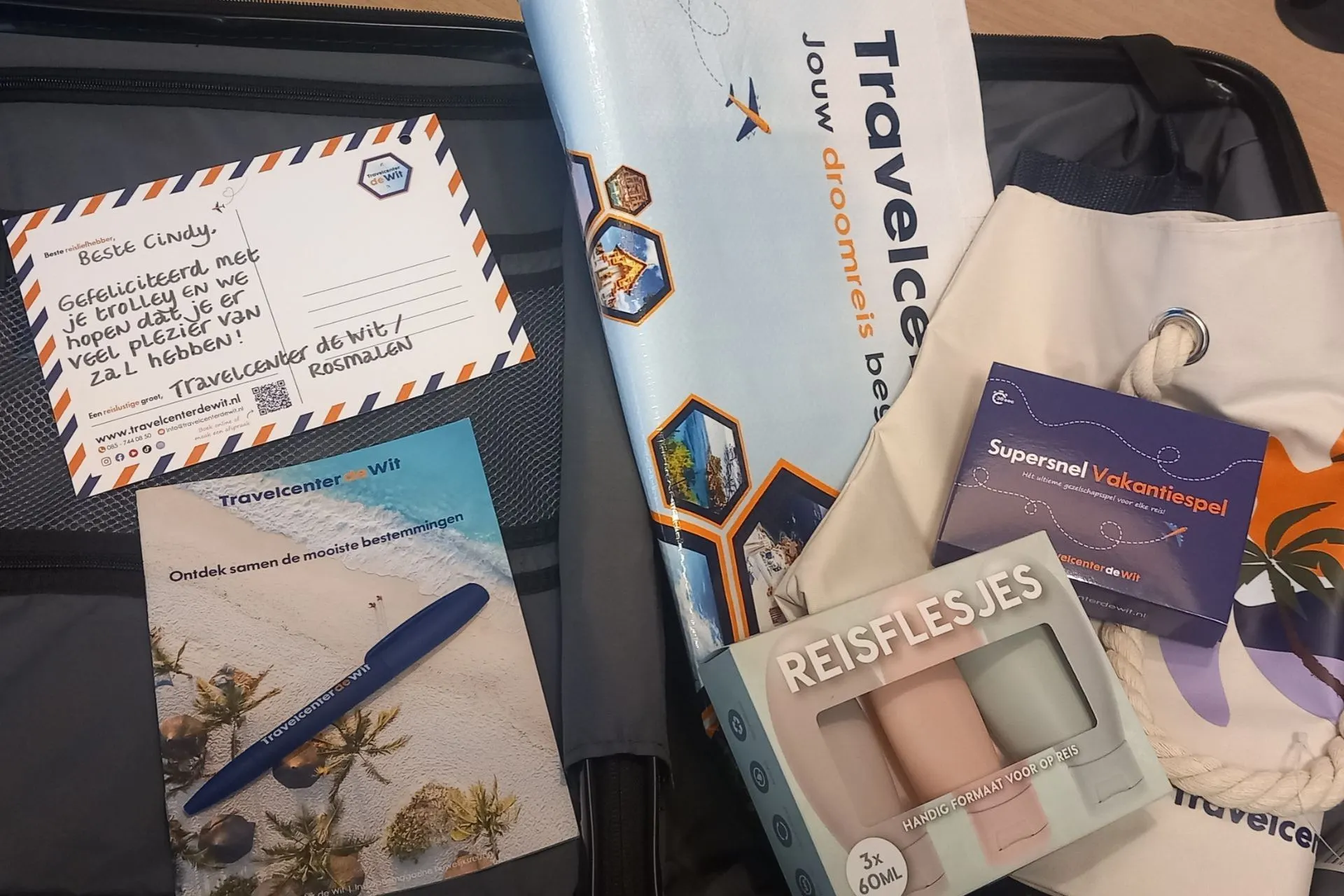 Een koffer gevuld met reisitems van Travelcenter de Wit, waaronder een ansichtkaart voor de winnaar, een pen, een brochure, een strandtas, reisflesjes en een reisspel. De prijs is samengesteld als reispakket.