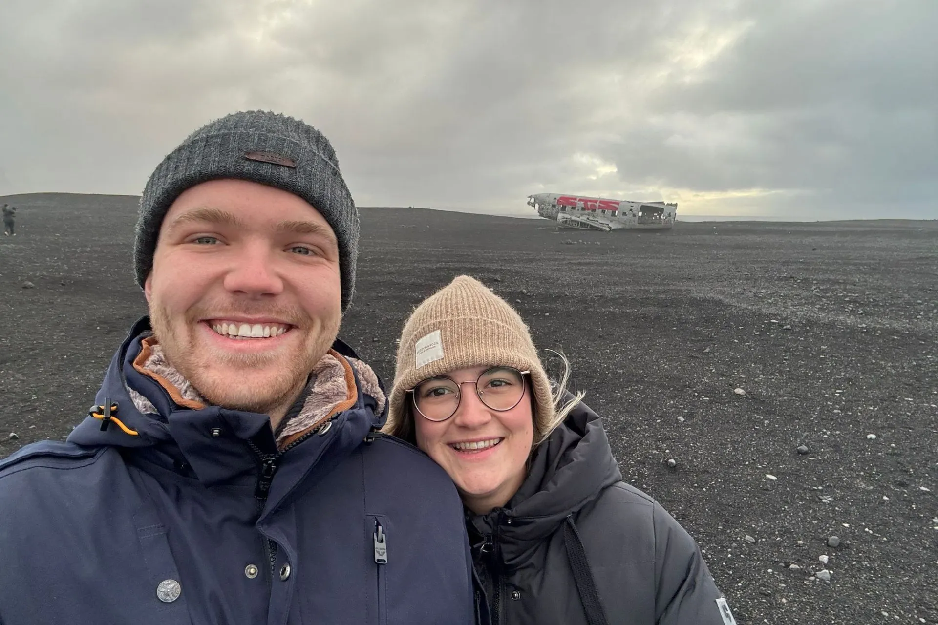 Reisadviseur Vera en haar vriend poseren op een zwart zandstrand in Reykjanes, IJsland, met op de achtergrond het wrak van het vliegtuig op het strand.