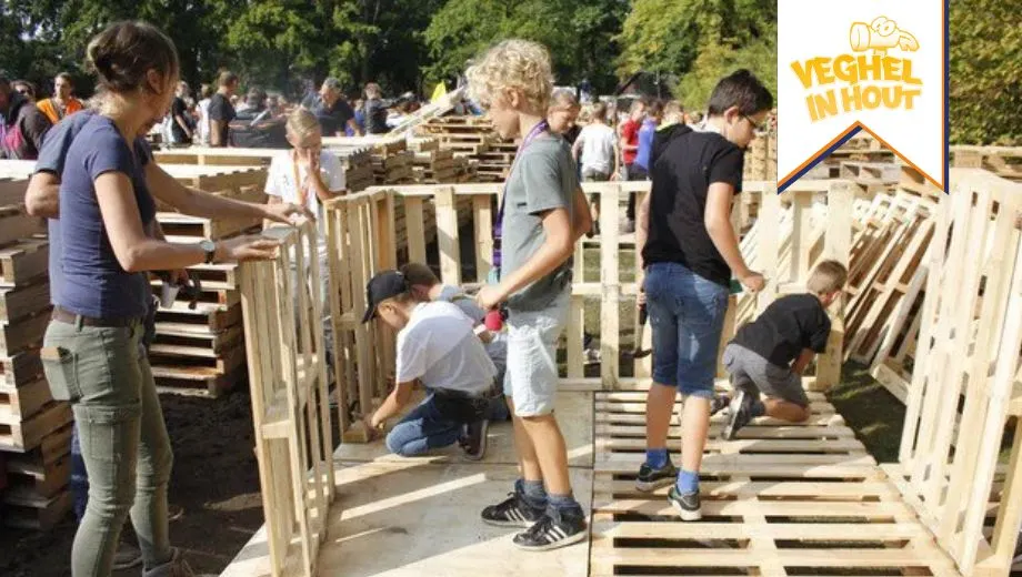 Kinderen bouwen samen houten hutten met pallets tijdens de kindervakantieweek ‘Veghel in Hout’. Op de achtergrond staan ouders en vrijwilligers toe te kijken. Rechtsboven is het logo van Veghel in Hout zichtbaar.