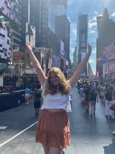 Een blonde vrouw staat in Times Square met haar handen in de lucht, omringd door de felle lichten van de enorme billboards en de levendige sfeer van de stad.