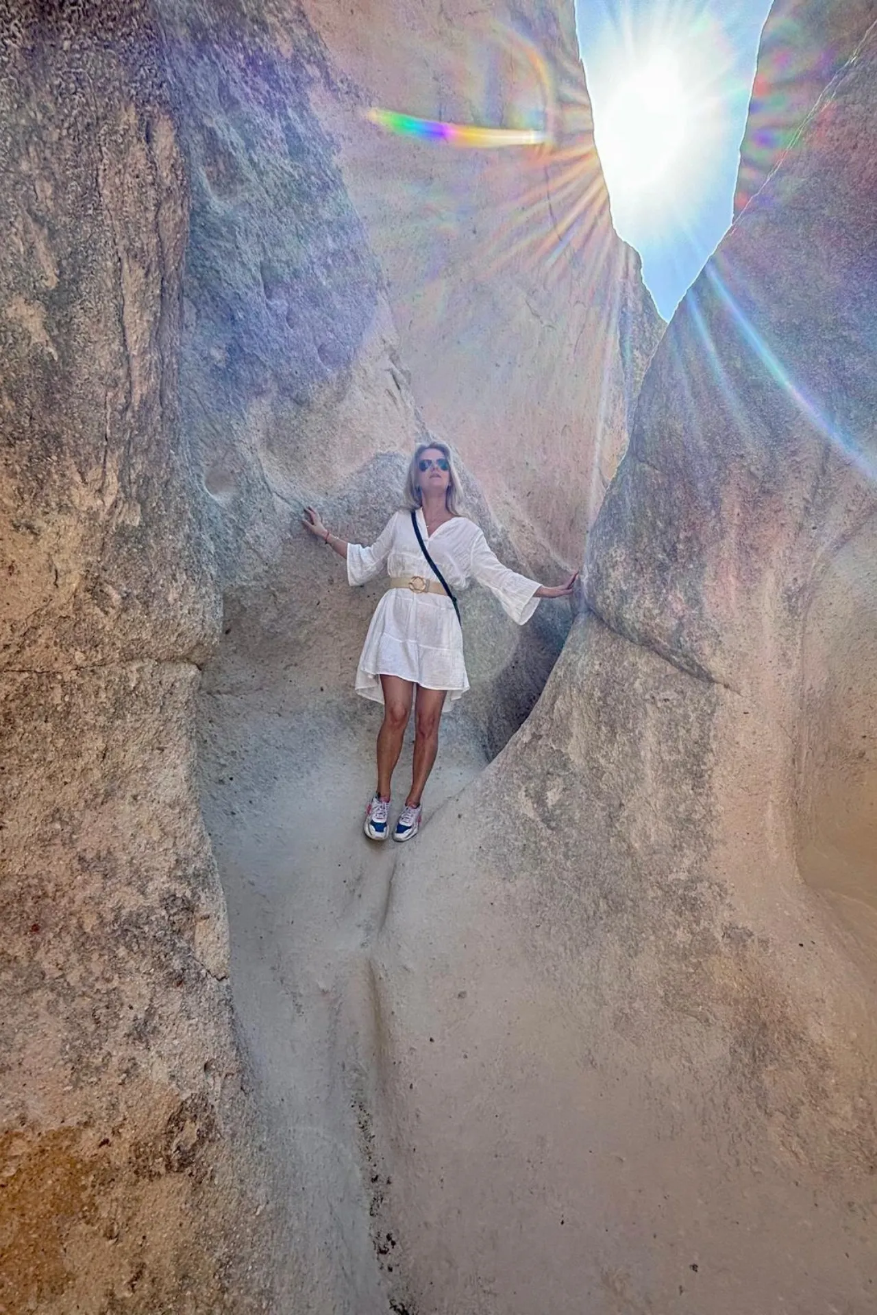 Persoon staat tussen hoge, smalle rotsformaties in Cappadocië, Turkije, gekleed in een lichte outfit die contrasteert met de aardetinten van het vulkanische gesteente. Zonlicht valt van boven in de kloof en creëert een prachtige lensflare, waardoor de scène een bijna magische uitstraling krijgt. De gladde rotswanden tonen sporen van natuurlijke erosie, typerend voor deze iconische regio. Deze locatie is ideaal voor avontuurlijke reizigers en fotografen die op zoek zijn naar unieke landschappen en bijzondere natuurwonderen in Anatolië.