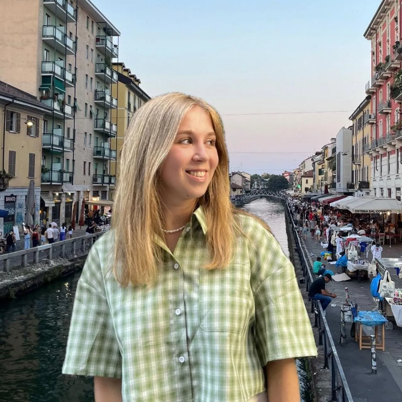 Marketeer Martje poseert lachend aan het kanaal van Navigli in Milaan, met op de achtergrond kleurrijke gebouwen, terrasjes en een levendige avondmarkt langs het water.