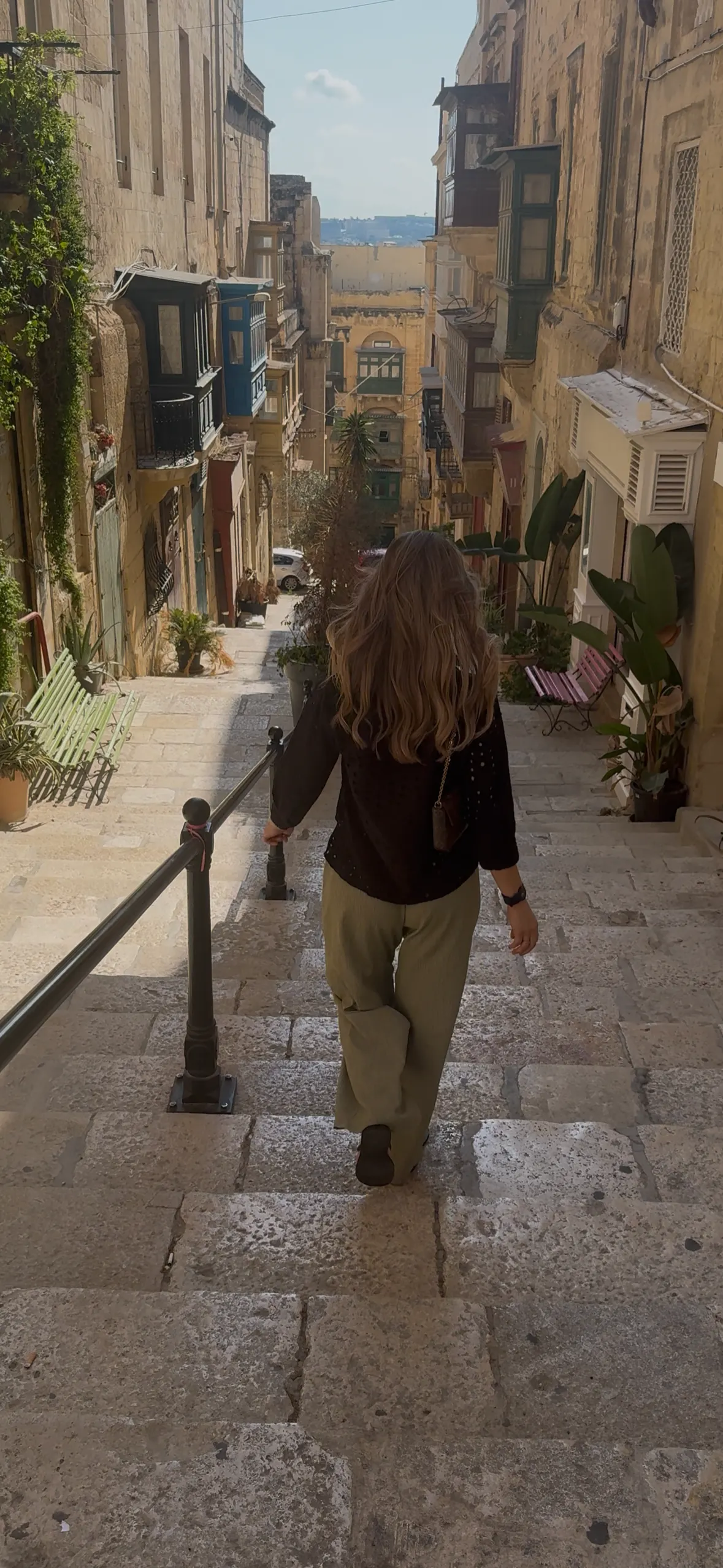 Emmie loopt door een smal straatje in Valetta Malta, gefotografeerd van achteren, met de charmante architectuur en het bruisende straatleven op de achtergrond.