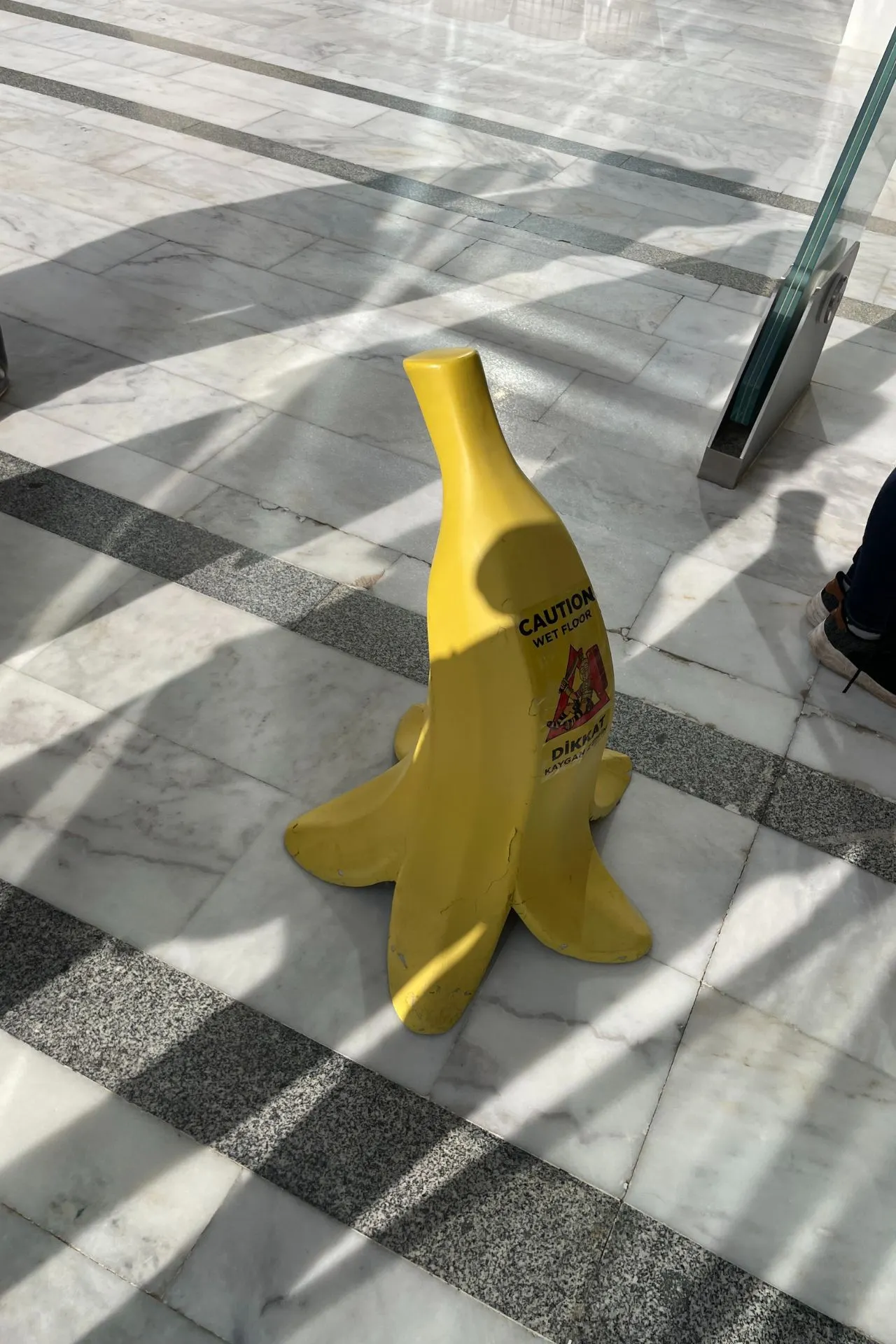 Gele decoratieve bananenschil op een marmeren vloer met de tekst “Caution Wet Floor”, gebruikt als grappige veiligheidswaarschuwing.