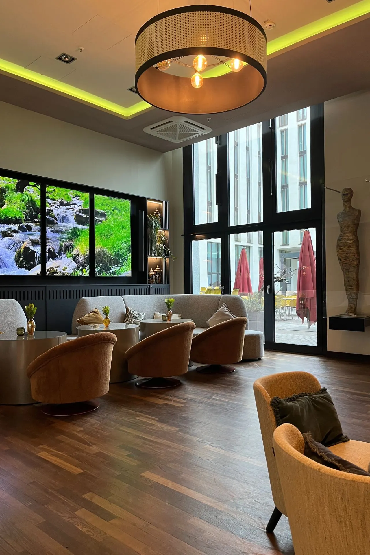 Een stijlvolle hotellounge met comfortabele stoelen, grote ramen met uitzicht op het terras en een warme, moderne inrichting. Leonardo Royal Munich Hotel. 