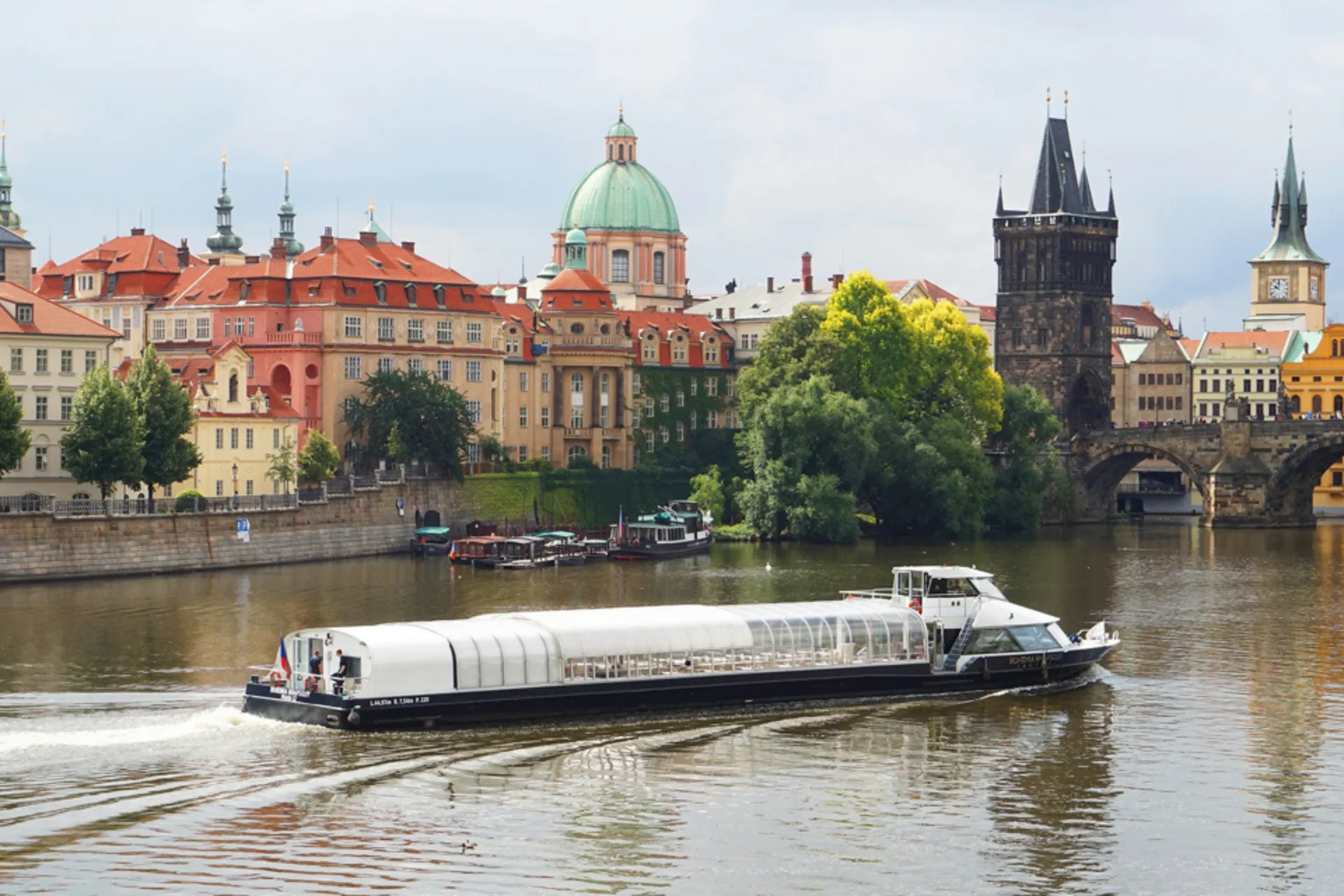 Rivier de Moldau met moderne rondvaartboot voor historische skyline van Praag, inclusief Karelsbrug, torens en kleurrijke barokgebouwen onder een bewolkte hemel.