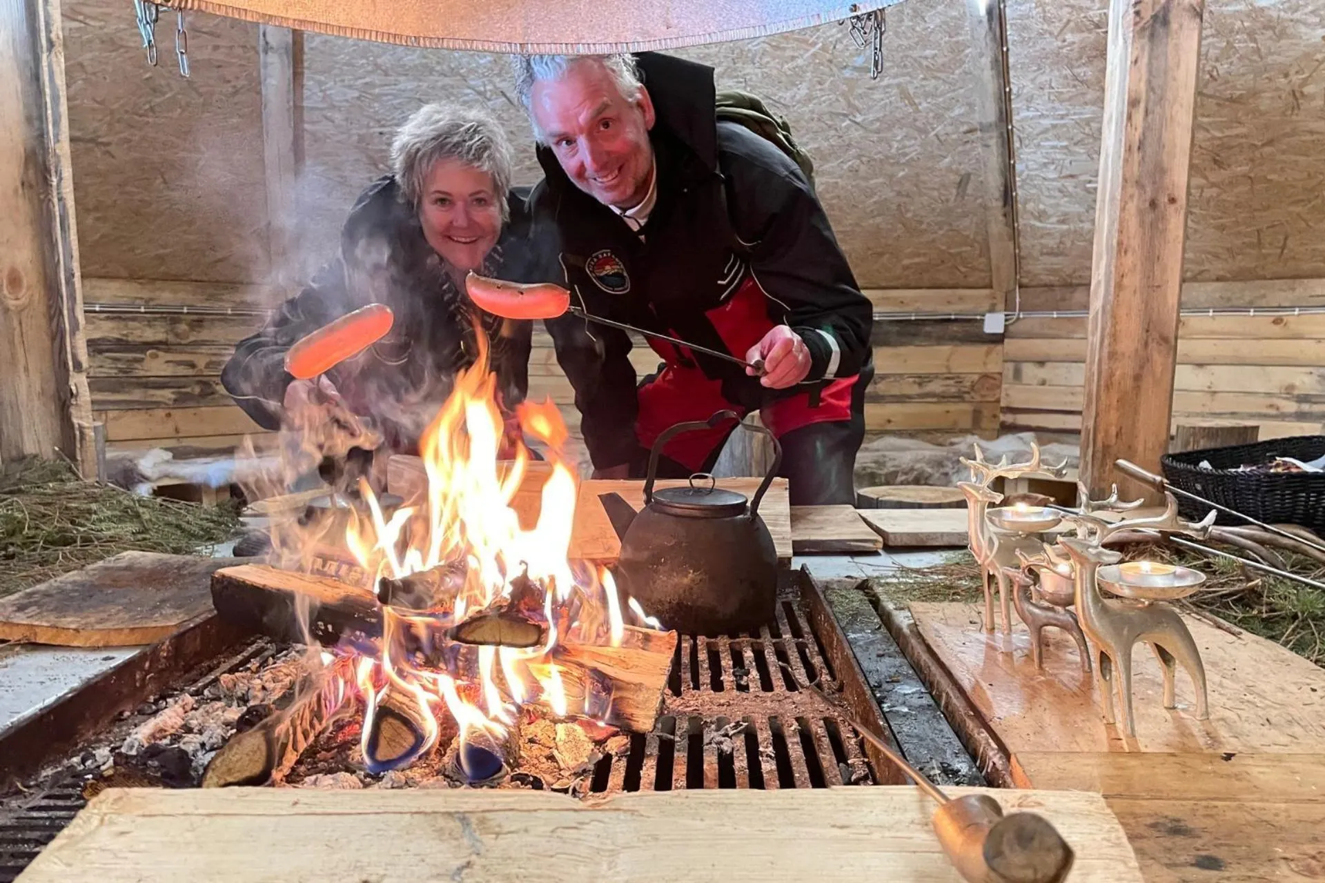Twee mensen roosteren worstjes boven een kampvuur in een traditionele hut in Fins Lapland

