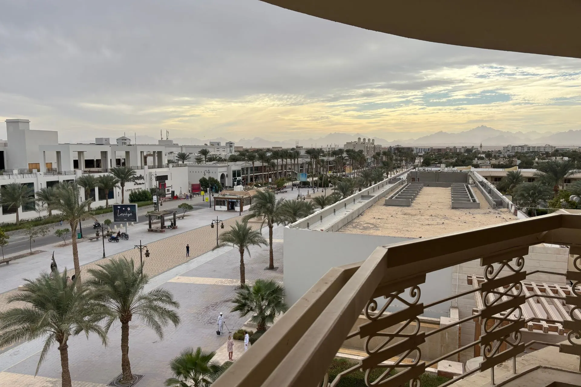 Uitzicht vanaf een balkon over een modern resort met witte gebouwen, palmbomen en een brede promenade. Op de achtergrond zijn bergen zichtbaar onder een bewolkte lucht met een lichte zonsondergang, wat een warme en serene sfeer geeft. Dit beeld benadrukt luxe en ruimte, ideaal voor promotie van zonvakanties, resorts en ontspanning in Egypte.