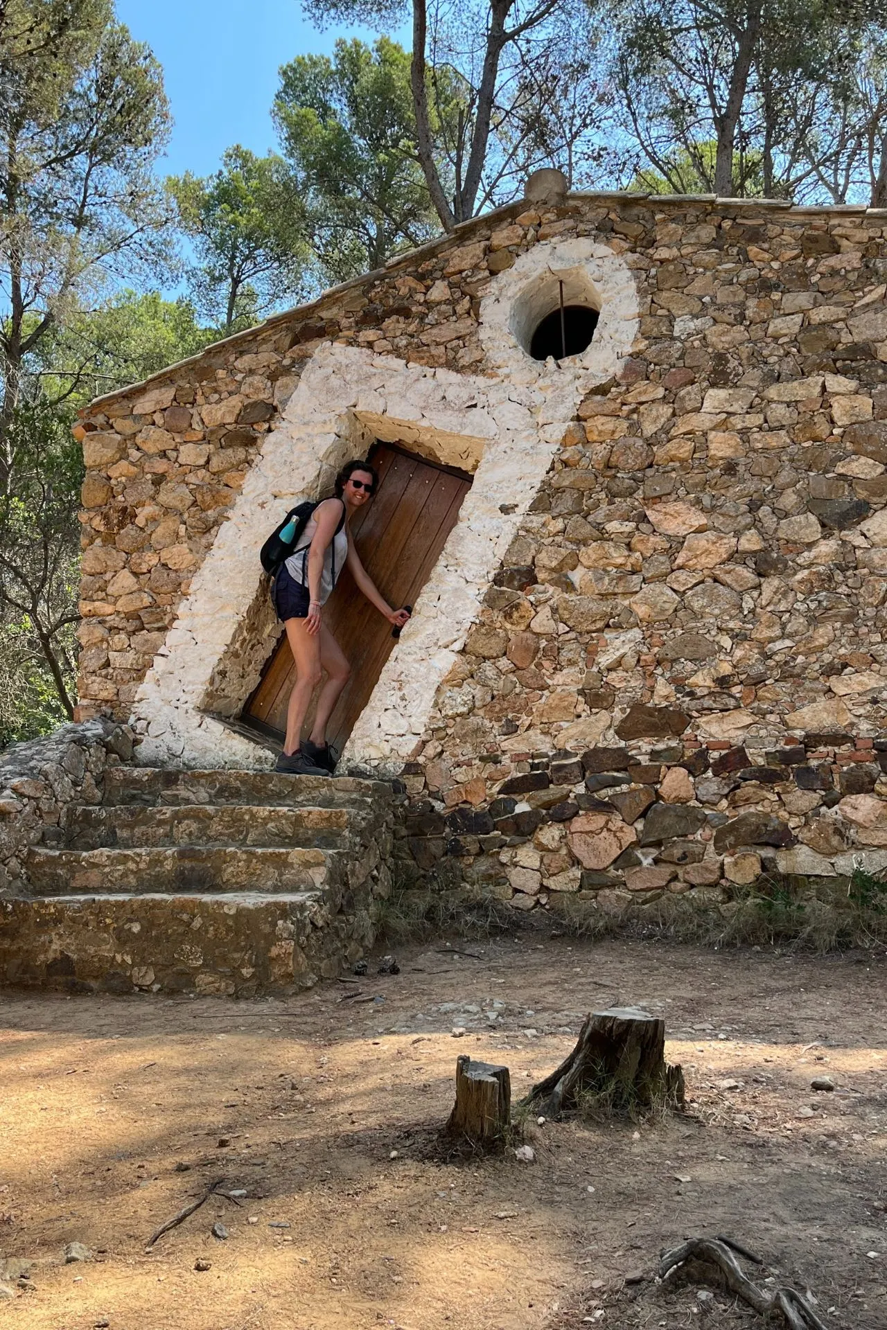 Stenen gebouw met een houten deur en witte omlijsting, gelegen in een bosrijke omgeving. Voor het gebouw staat een persoon met een rugzak in de deuropening, omringd door bomen en zonverlichte grond.