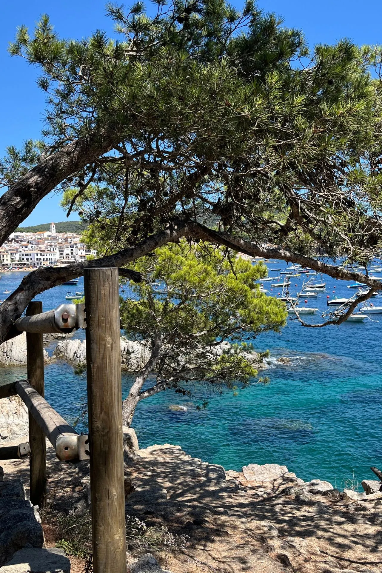 Uitzicht vanaf een rotsachtig pad met houten railing, geflankeerd door pijnbomen. Op de achtergrond ligt een helderblauwe zee met meerdere kleine boten en een kustlijn met witte gebouwen onder een strakke blauwe lucht.