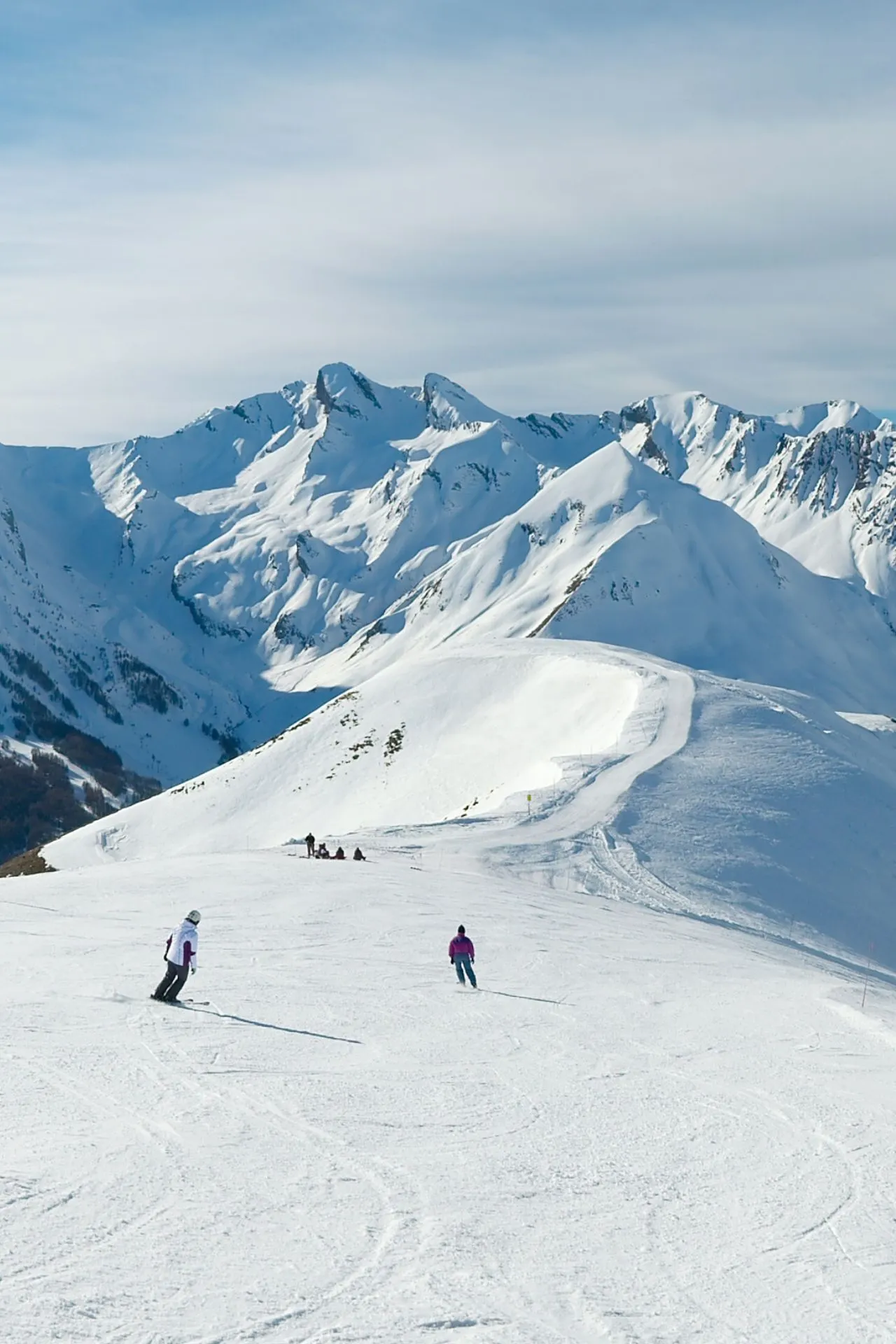 Skiërs dalen af op een Italiaanse skipiste met op de achtergrond majestueuze, besneeuwde bergtoppen en een helderblauwe winterlucht.