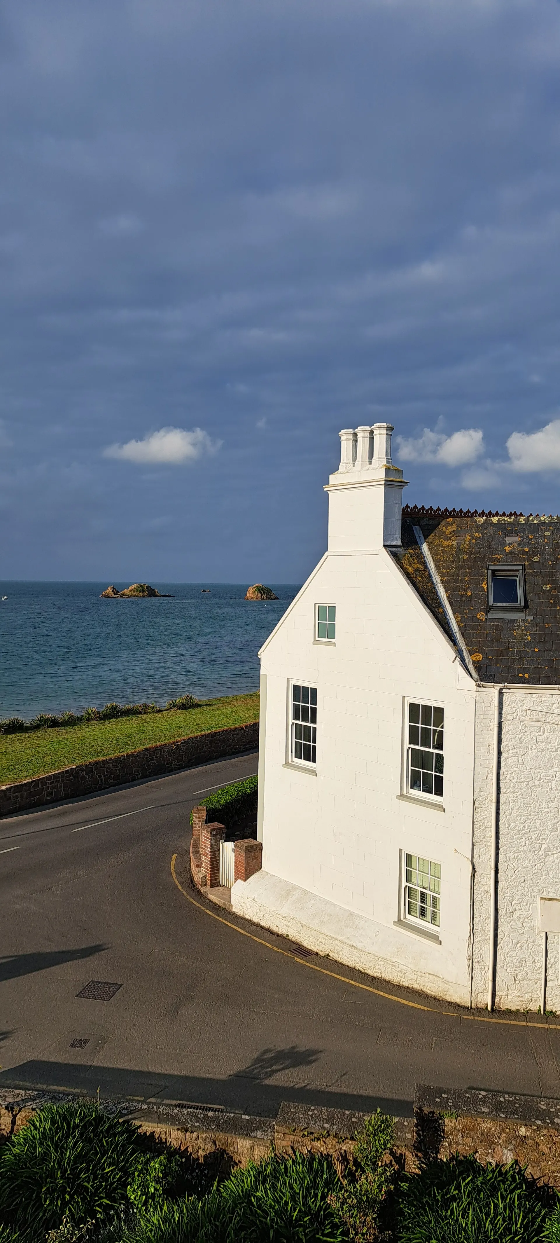 Traditioneel wit huis met grijze dakpannen gelegen aan een bochtige kustweg, met uitzicht op helderblauw zeewater en rotsformaties in de verte. De woning heeft meerdere ramen en een schoorsteen, omringd door groene struiken en een stenen muur langs de weg. Perfect voor kustvakanties, pittoreske accommodaties en fotografie van charmante architectuur met panoramisch uitzicht op zee en een rustige omgeving.

