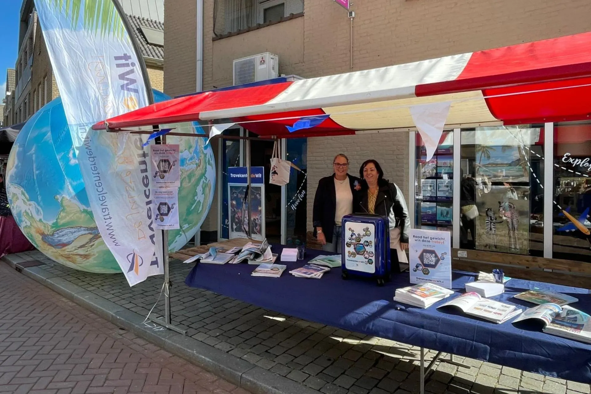 Twee medewerkers van Travelcenter de Wit staan lachend achter een marktkraam tijdens het KOM Festival in Rosmalen. De kraam is versierd met vlaggetjes en brochures over reizen. Op de achtergrond is een grote opblaasbare wereldbol te zien.