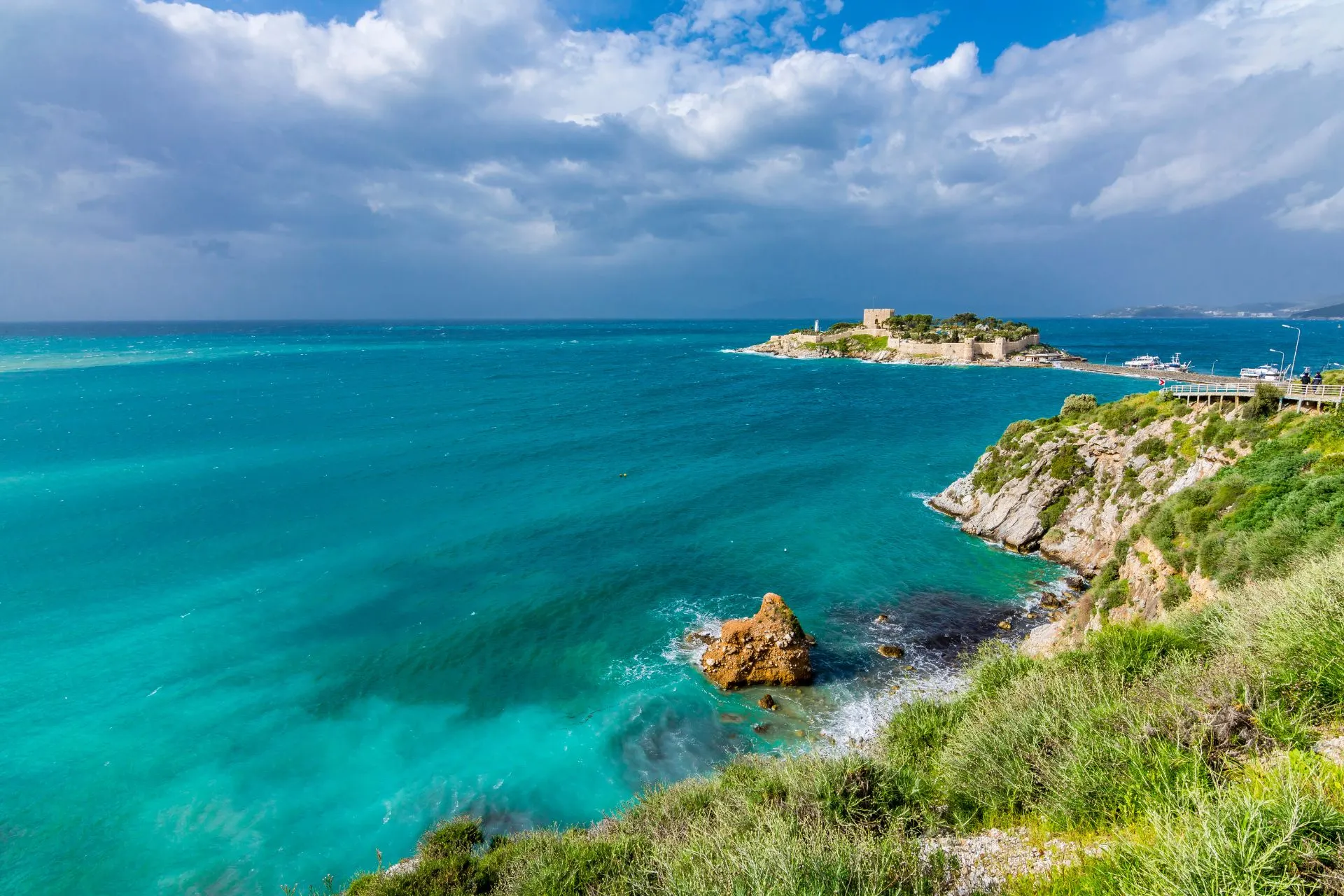 Uitzicht op de turquoise zee en het Duiveneiland (Güvercinada) voor de kust van Kuşadası, met een historisch fort en wandelpad.