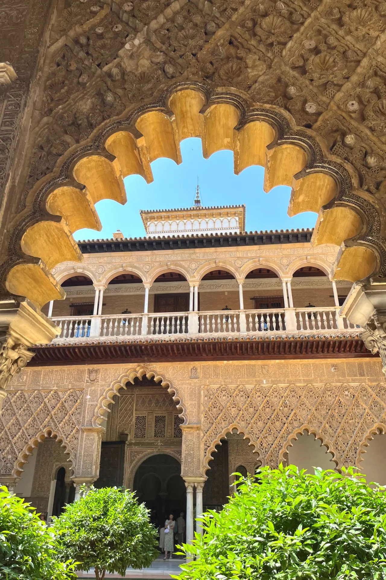 Binnenhof van het Real Alcázar in Sevilla met een prachtig uitzicht door een rijk versierde hoefijzervormige boog. De gevel toont Moorse architectuur met sierlijke patronen, bogen en een balkon met zuilen. Voorin staan sinaasappelboompjes in de zon.
