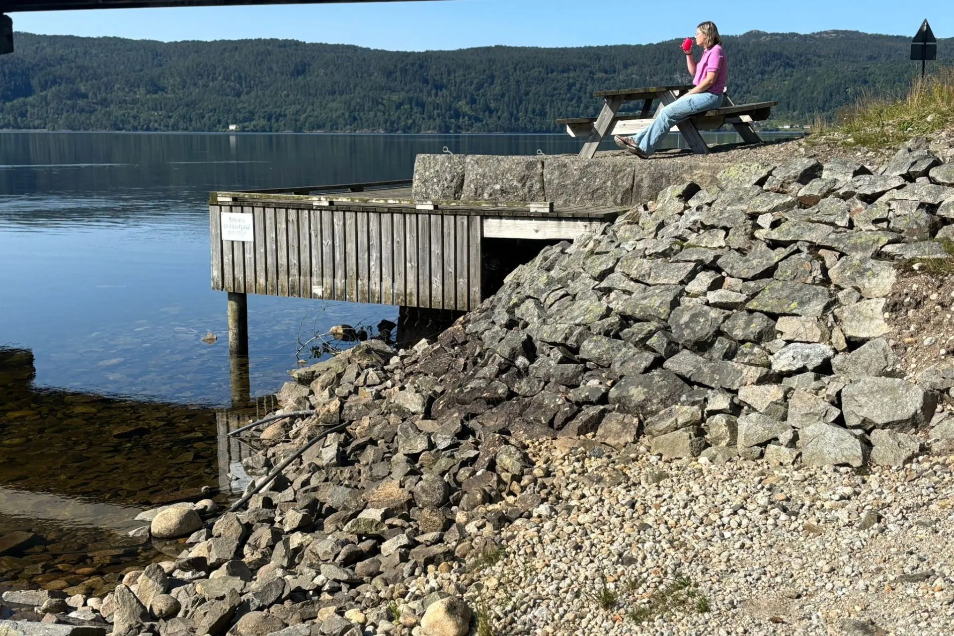 Persoon zit op een houten steiger aan een meer, drinkt uit een beker en kijkt uit over het water. Op de voorgrond liggen grote stenen en grind, op de achtergrond beboste heuvels onder een heldere blauwe lucht.