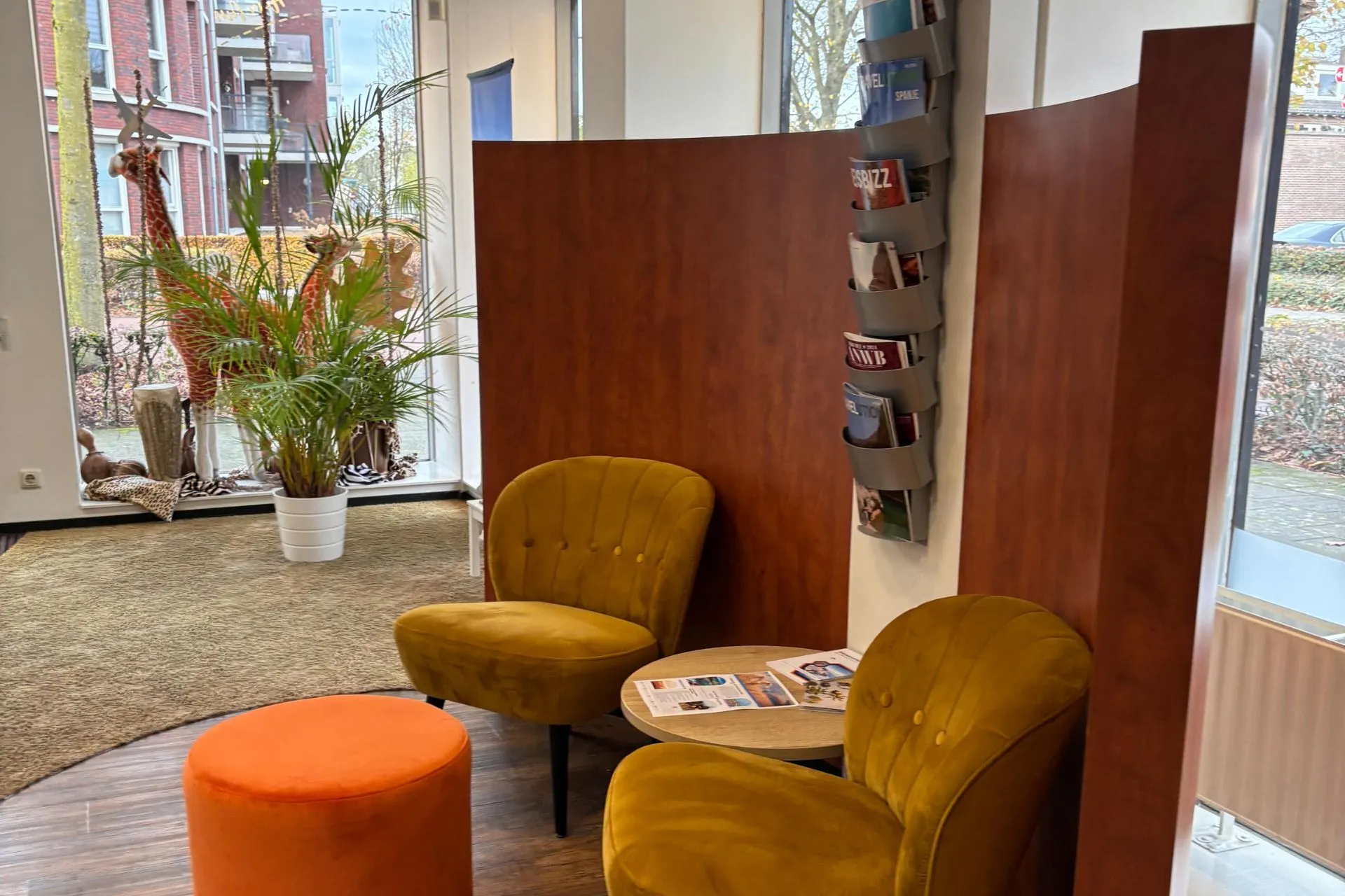 Ontvangstruimte van Travelcenter de Wit in Heesch. Geel fluwelen stoelen en een oranje poef rond een lage tafel met reisbrochures. Aan de wand een rek met tijdschriften. Grote ramen met uitzicht op buiten, aangekleed met planten en decoratieve giraffen.