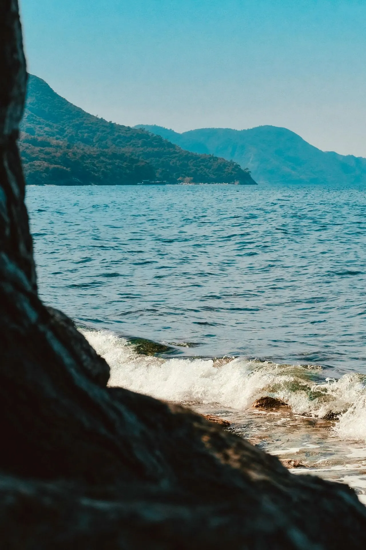 Langs een rustige kustlijn slaan kleine golven tegen de rotsen, terwijl in de verte groene heuvels oprijzen boven het helderblauwe water. Deze serene omgeving roept het gevoel op van ontspannen bestemmingen die perfect zijn voor een familie cruise, waar gezinnen samen kunnen genieten van natuur en zee. De kalmte van het landschap maakt het ideaal voor een gezinscruise waarbij iedereen tot rust kan komen en de omgeving op eigen tempo ontdekt.