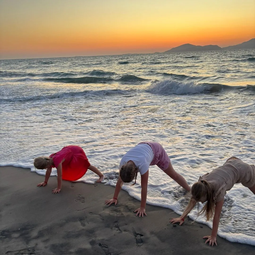 Drie kinderen buigen zich lachend over het zand terwijl de golven hun voeten raken, met een kleurrijke avondlucht op de achtergrond
