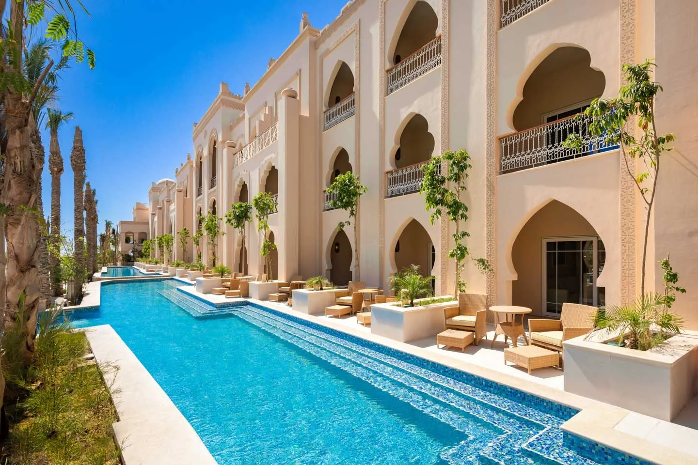 Luxe resort The Grand Palace in Hurghada, Egypte, met een elegant zwembad dat langs een rij kamers en suites loopt. De architectuur heeft Arabische invloeden met grote bogen, sierlijke details en lichte zandkleurige gevels. Rond het zwembad staan comfortabele ligstoelen en tafels, omgeven door groene planten en palmbomen, wat een tropische en ontspannen sfeer creëert. Perfecte bestemming voor een luxe zonvakantie, ontspanning en genieten van het warme klimaat aan de Rode Zee.