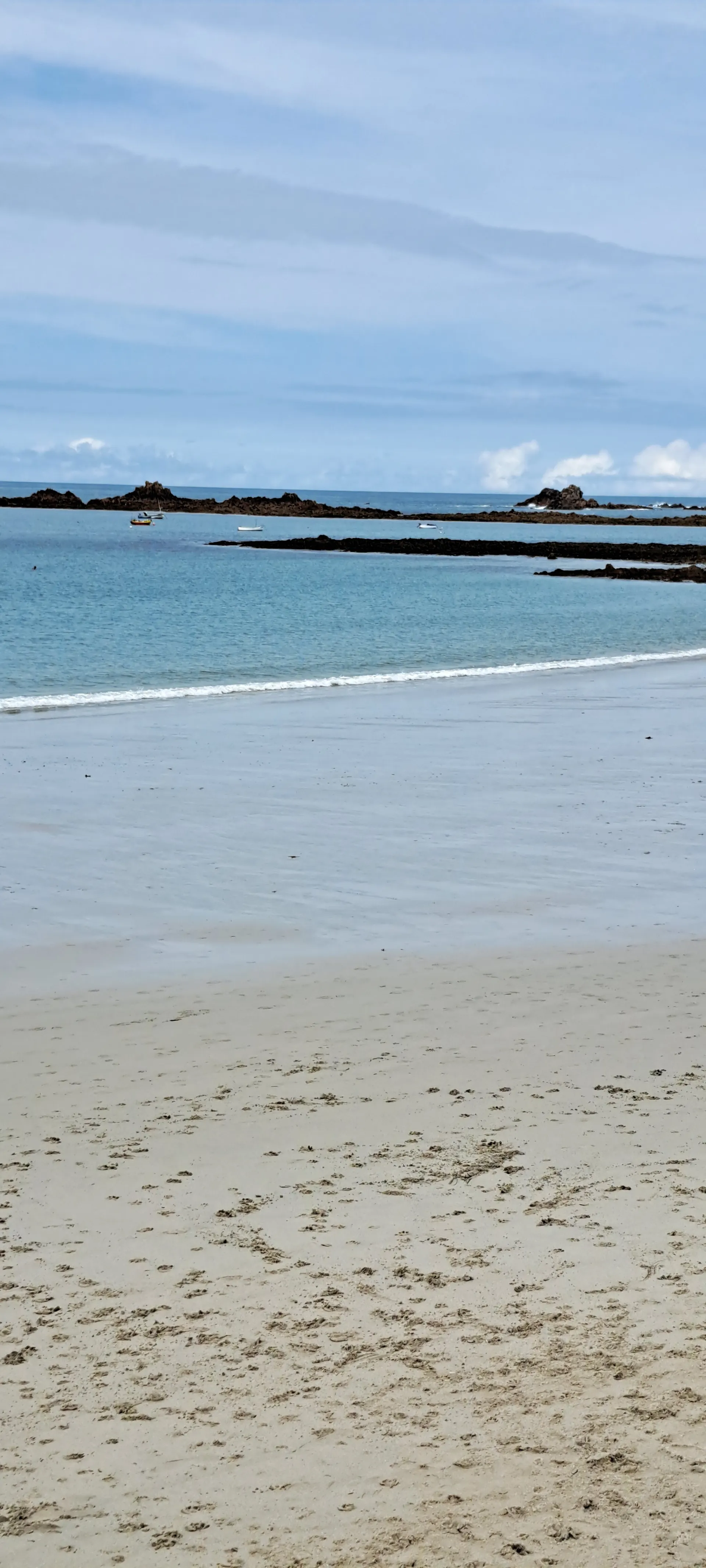 Uitgestrekt zandstrand met fijn licht zand dat overgaat in helderblauw zeewater, omringd door donkere rotsformaties die uit de kustlijn steken. De rustige branding en het kalme water creëren een serene sfeer, ideaal voor strandwandelingen, zonnebaden en fotografie. Deze schilderachtige kustlocatie biedt een prachtig uitzicht op de oceaan en is perfect voor toeristen die op zoek zijn naar ontspanning, natuur en ongerepte stranden langs een idyllische kustlijn.