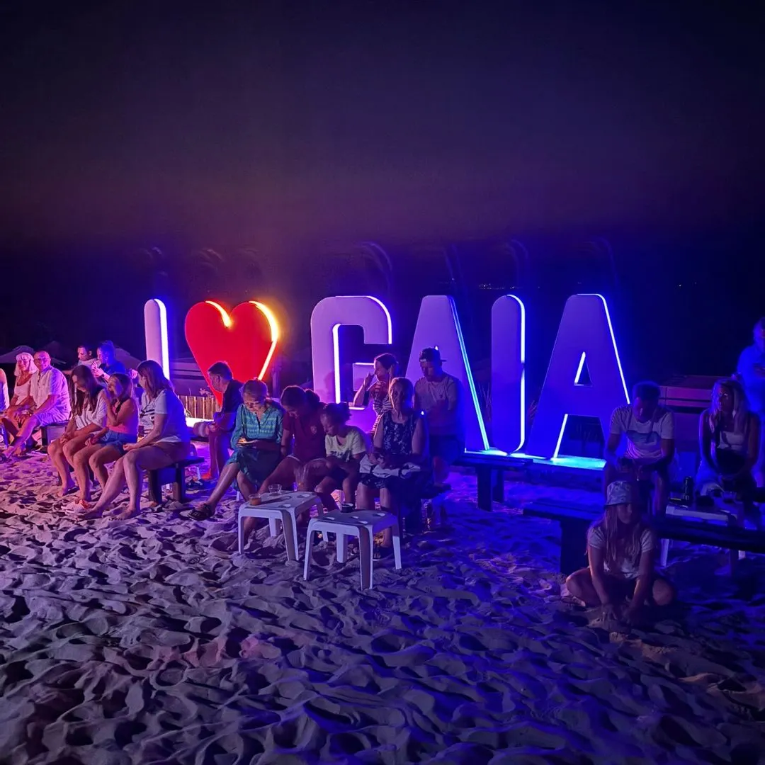 Gezellige avondsfeer op het strand van het Gaia Royal Hotel, met kleurrijk verlichte “I ❤️ GAIA” letters en families op banken in het zand