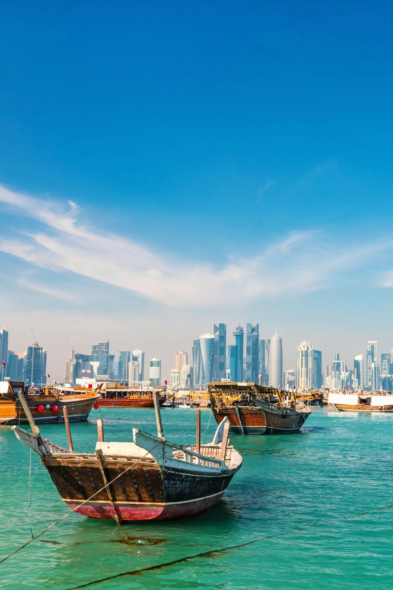 Traditionele houten dhows dobberen op het turquoise water voor de moderne skyline van Doha, Qatar, met indrukwekkende wolkenkrabbers onder een heldere blauwe lucht.