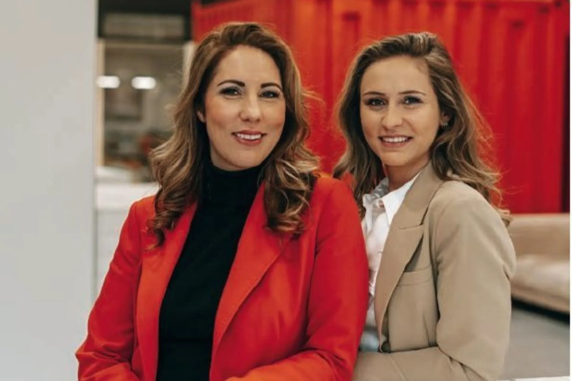 Twee professioneel geklede vrouwen staan naast elkaar in een moderne werkomgeving met een warme, rode achtergrond. Beiden dragen een stijlvolle blazer, één in rood en één in beige, gecombineerd met een nette top en blouse. De setting straalt een zakelijke en professionele sfeer uit, passend bij thema’s als ondernemerschap, vrouwelijke professionals, teamwerk en corporate branding. Deze afbeelding is geschikt voor gebruik op websites over marketing, consultancy, zakelijke dienstverlening, vrouwen in het bedrijfsleven en professionele fotografie voor bedrijven.