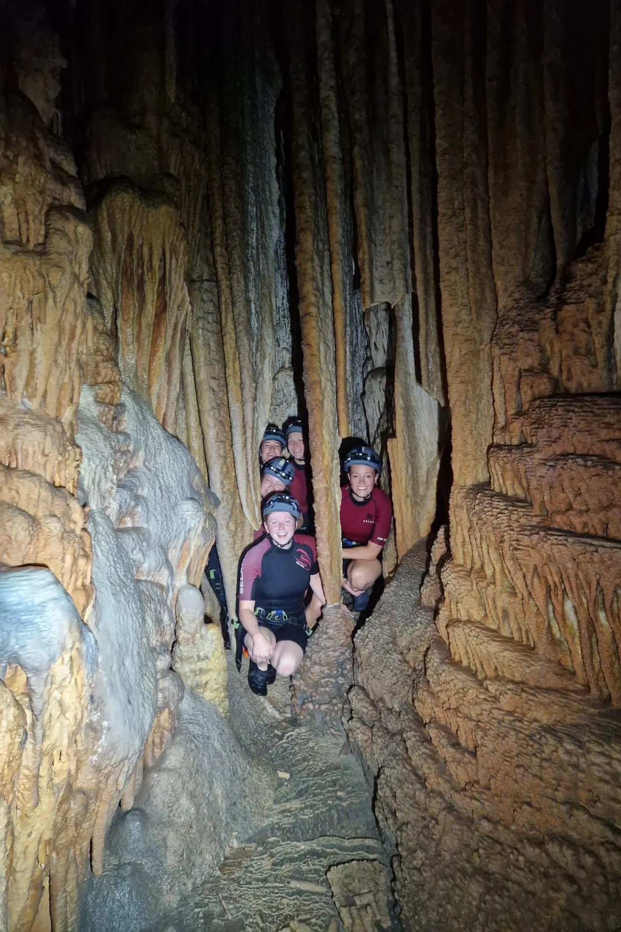 Groep mensen in wetsuits en helmen kruipt door een smalle doorgang tussen indrukwekkende stalactieten en stalagmieten in een grot op Mallorca.