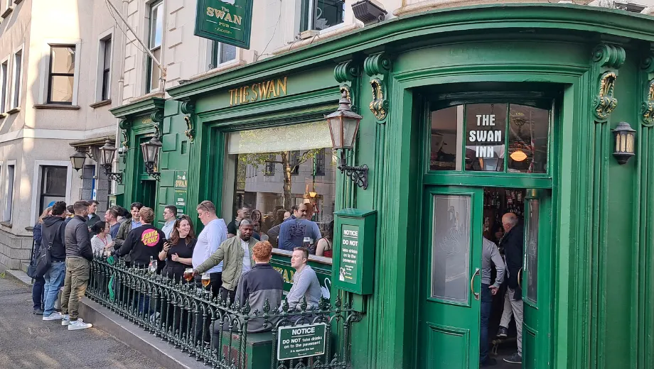 Traditionele Engelse pub genaamd “The Swan Inn” met een opvallende groene gevel, grote ramen en klassieke lantaarns aan de buitenkant. Voor de ingang staat een groep mensen gezellig samen, terwijl binnen meer gasten zichtbaar zijn. Het terras is omheind met een sierlijk hekwerk en er hangt een bord met informatie over het café. Deze locatie straalt authenticiteit en gezelligheid uit, ideaal voor liefhebbers van pubs, lokale cultuur en sociale ontmoetingen in een historische stadsomgeving.