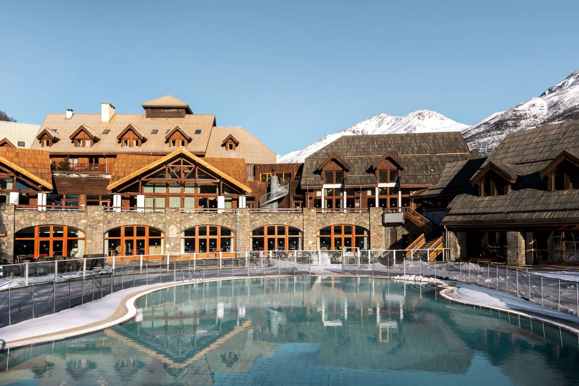 Buitenzwembad bij Club Med skiresort in de Alpen, omringd door houten chalets en besneeuwde bergen. Beleef een luxe all-inclusive wintersportvakantie met wellness, skipas en skilessen inbegrepen.