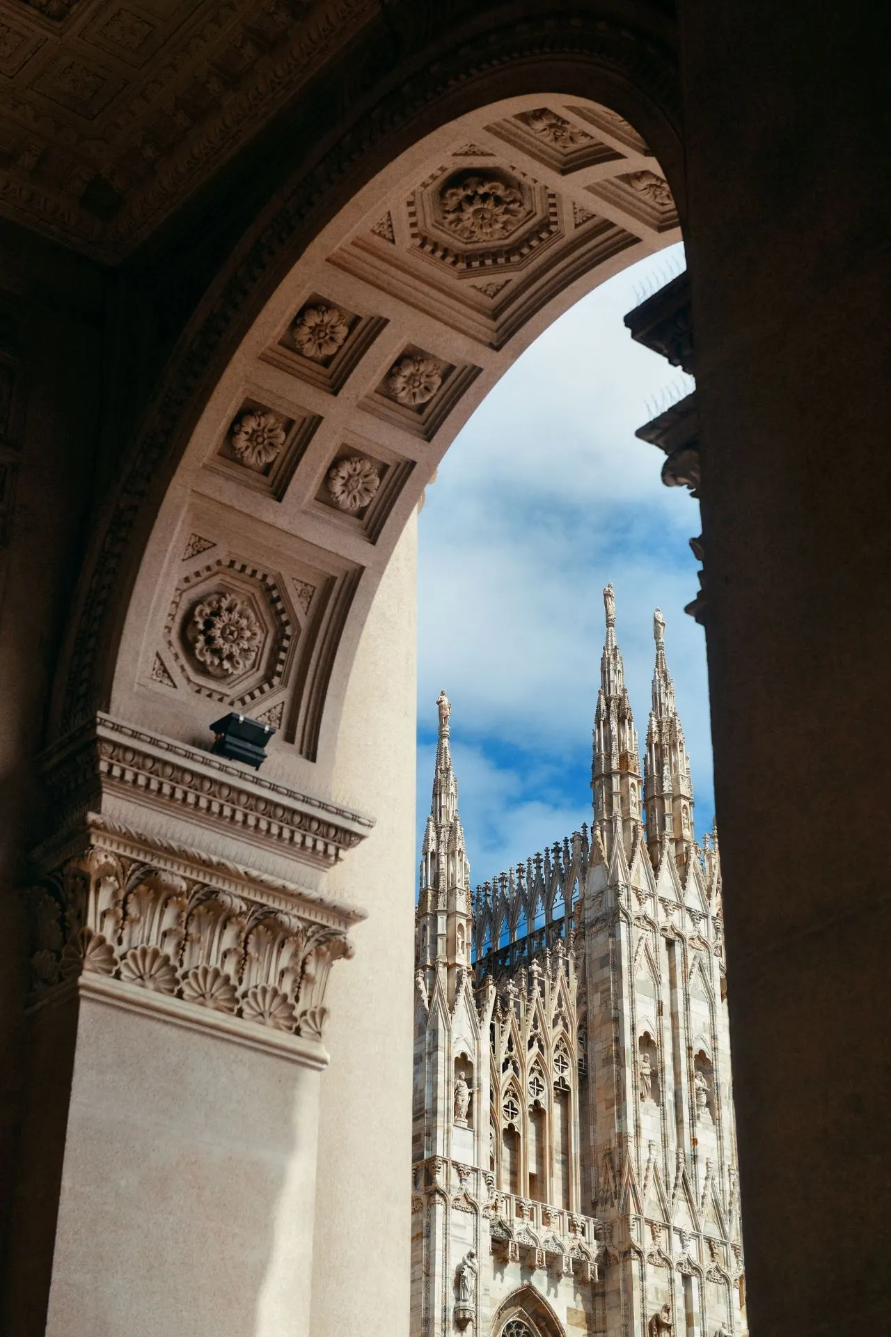 Zicht op de Duomo di Milano vanuit een monumentale boog met rijk versierde plafondpanelen. De gotische torens van de kathedraal steken omhoog tegen een blauwe lucht met wat wolken.